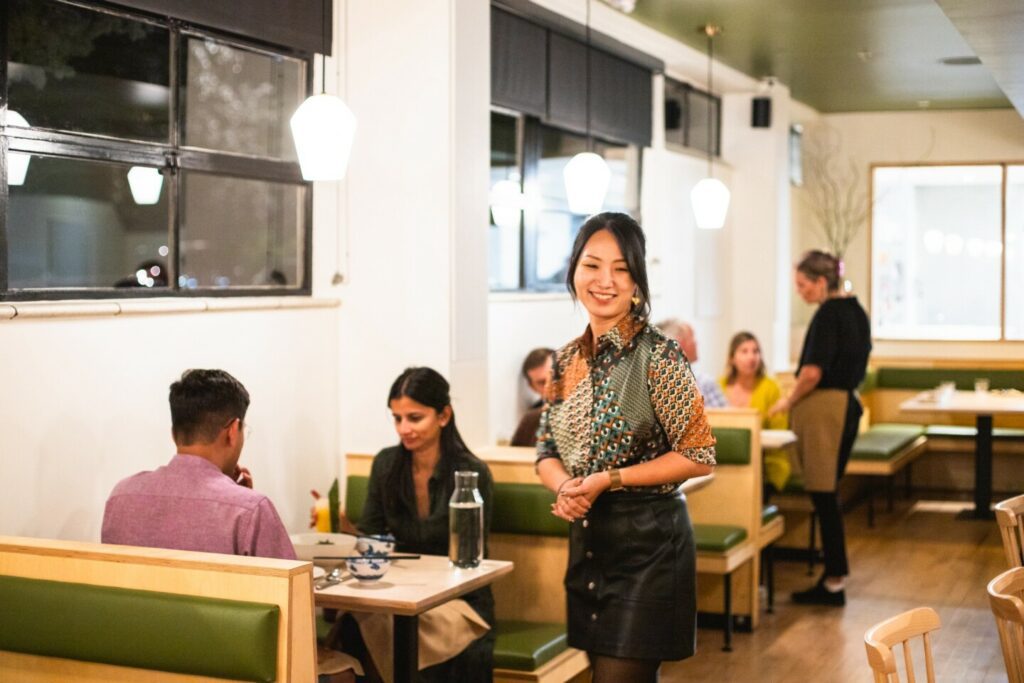 A woman standing in the middle of a restaurant's white dining room with several diners eating at booths