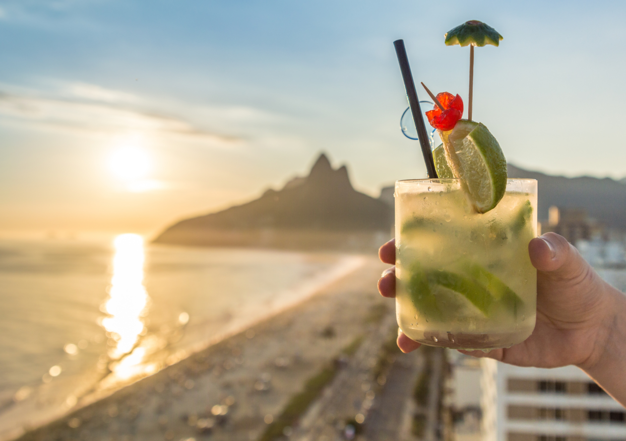 Hand Holding a Cachaça Cocktail, Selective Focus, Sunset on Beach in the Background