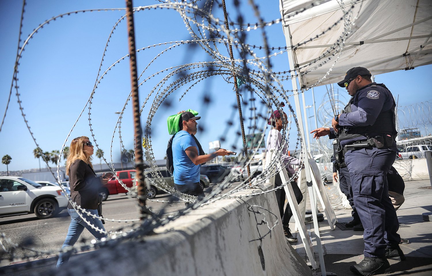 Three pedestrians are seen framed by razor wire as they step toward a border agent in uniform under a tent just past a low jersey barrier. Cars and palm trees are seen on the horizon.