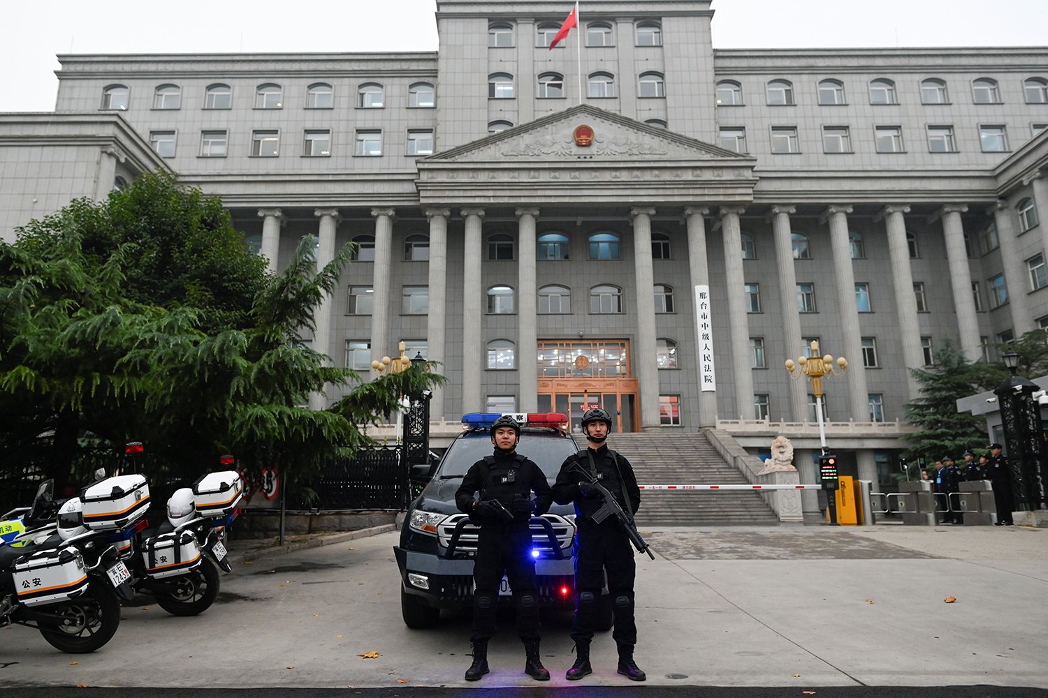 Two police officers holding guns stand in front of a police vehicle in front of a multi-story courthouse. At left are the tail ends of two motorcycles.