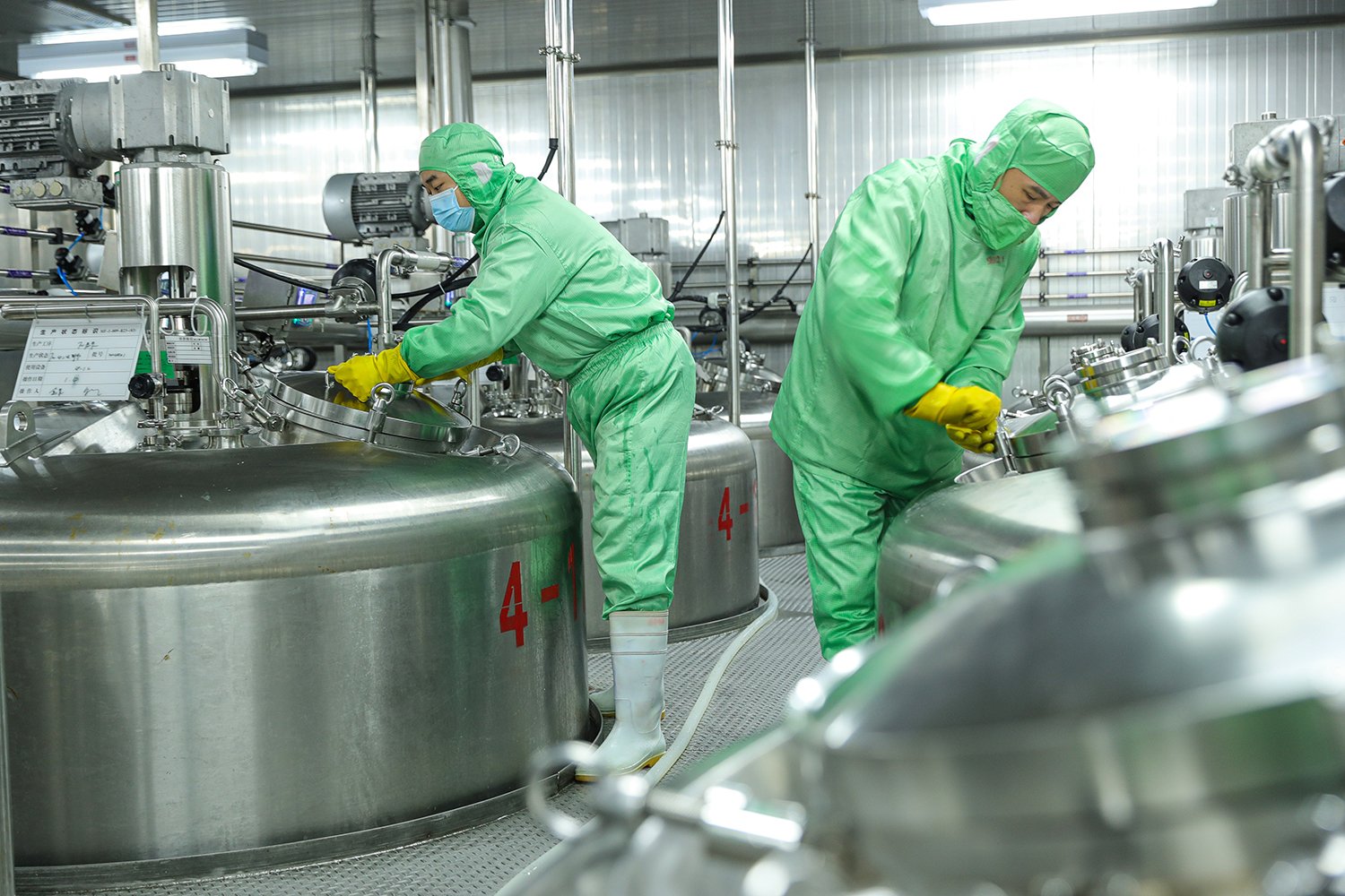 Two workers in protective suits, masks, and gloves, work alongside giant metal vats in a manufacturing facility.