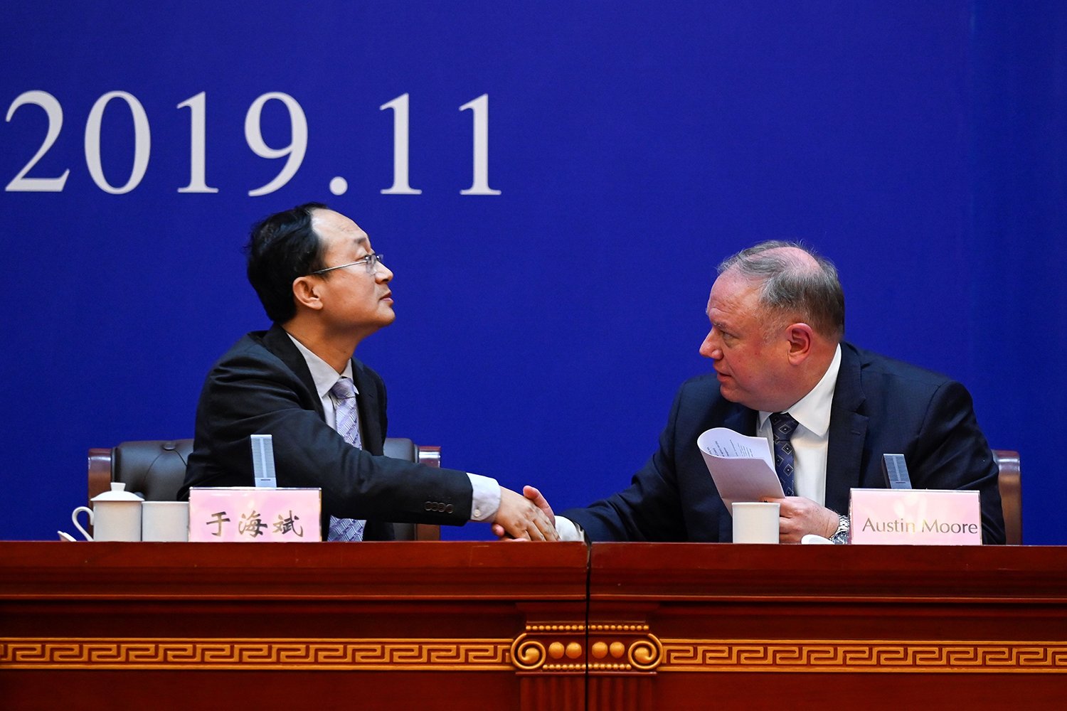 A Chinese man lifts his chin and holds out his hand for a handshake with an American man as they look toward each other. They sit behind a dais under a blue backdrop with the numbers 2019.11 on it.