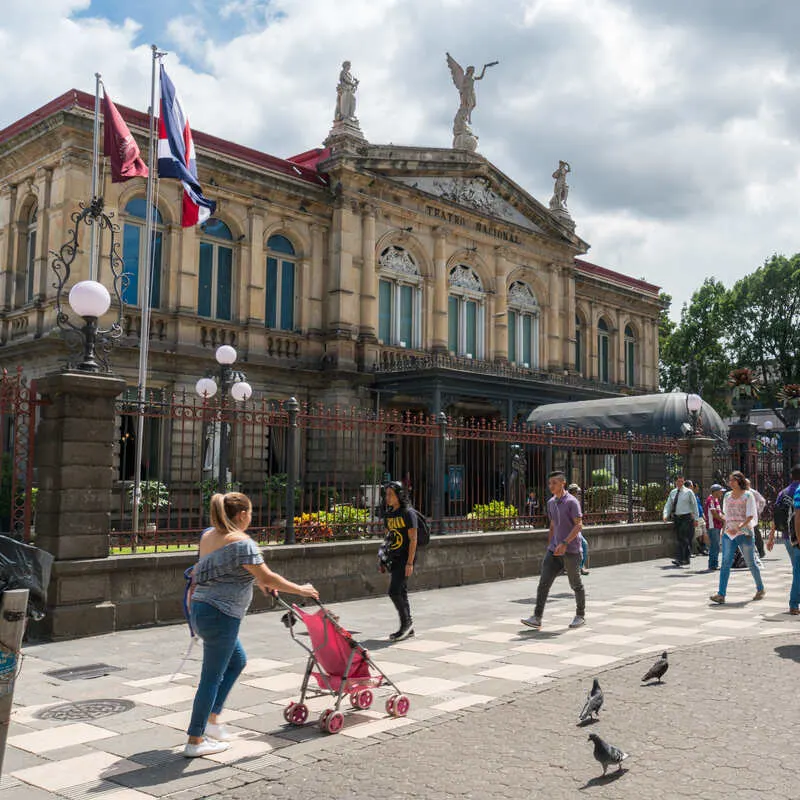 People Strolling Down Past A Colonial-Style Building In A Pedestrianized Street In San Jose, Capital City Of Costa Rica, Central America