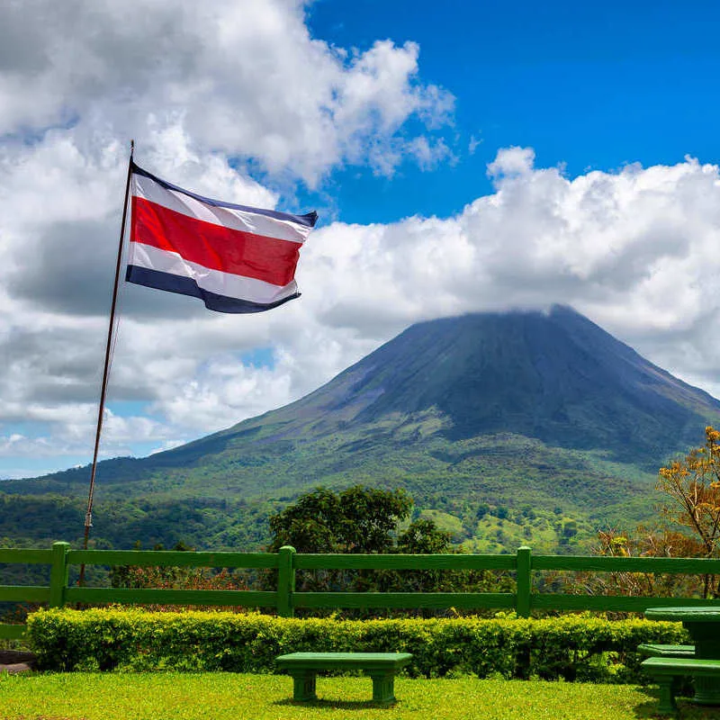 Costa Rican National Flag Flying On A Flagpole With The Arenal Volcano Shown In The Background, Covered By Clouds, Costa Rica, Central America