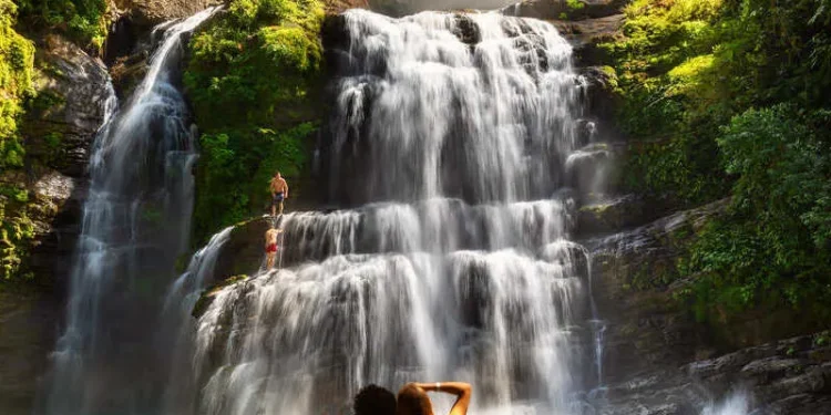Couple Of Travelers Sat On A Rock As They Admire A Waterfall In Manuel Antonio National Park In Costa Rica, Central America