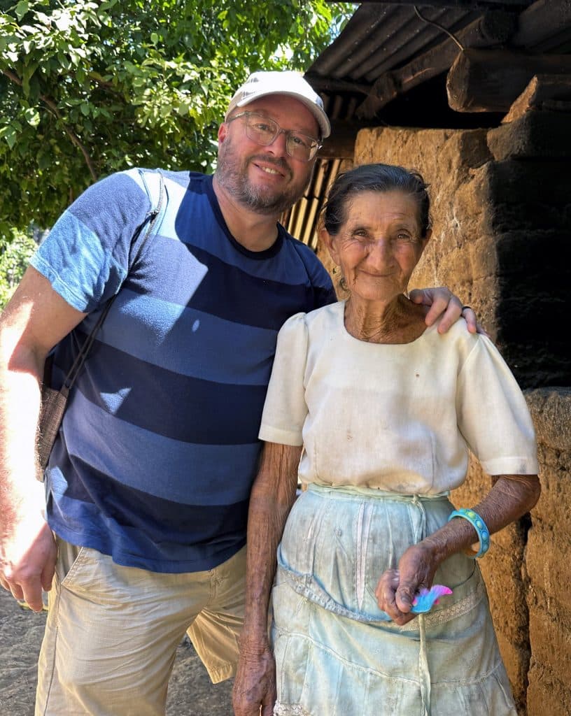 A man and an elderly woman stand together smiling. The man wears a blue striped shirt, beige pants, glasses, and a cap. The woman wears a white blouse, a light blue skirt, and holds a small colorful object. They are outside, with greenery and part of a rustic building visible in the background.