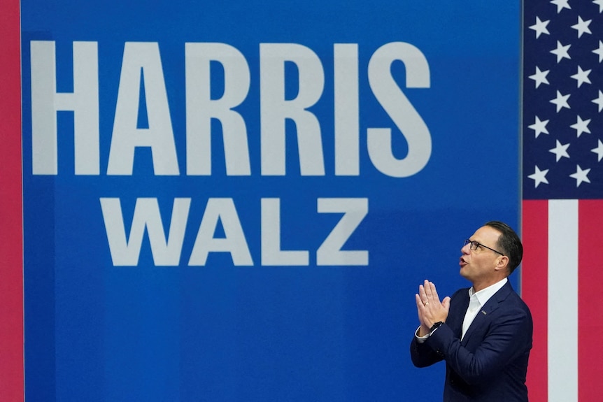 Josh Shapiro holds his hands together as he stands in front of the giant words 'Harris Walz' on stage.