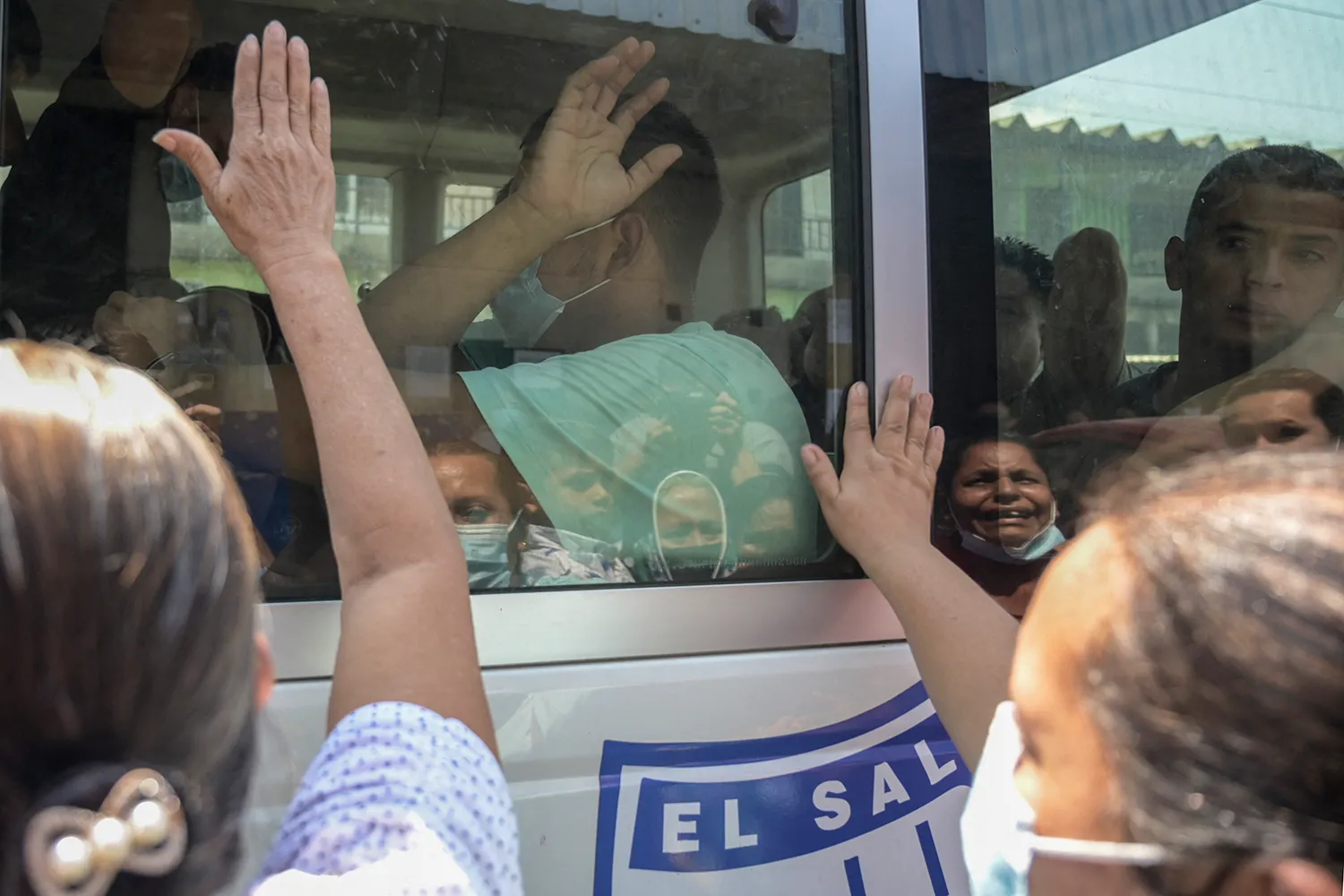 Prisoners are loaded onto a bus while mourning family members press their hands to the glass. from outside. One of the prisoners lifts his hand to wave to them.