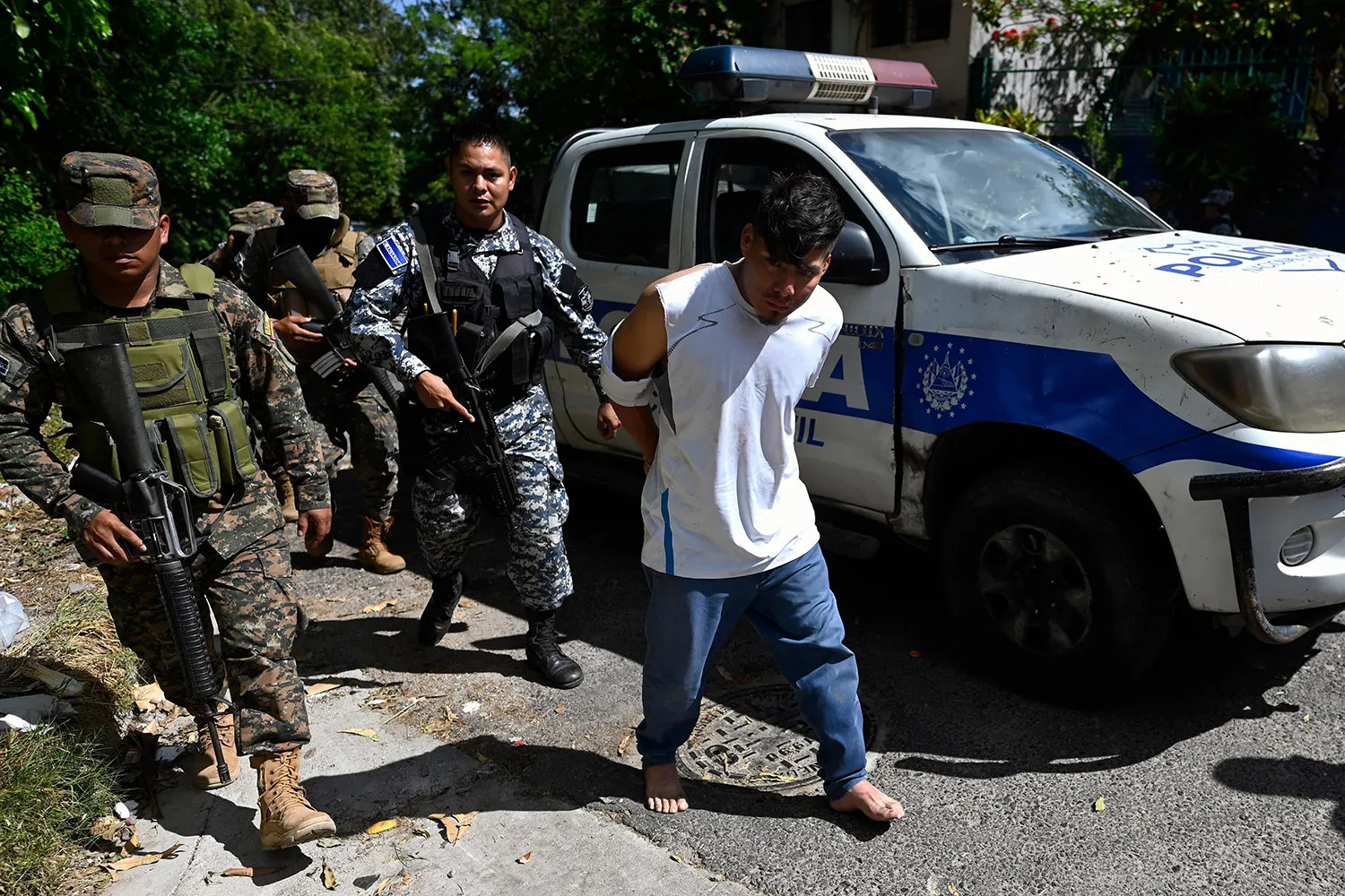 Police and soldiers escort people captured alongside a police vehicle during an operation against gang members. The detainee closest to the camera has his hands behind his back and bare feet.