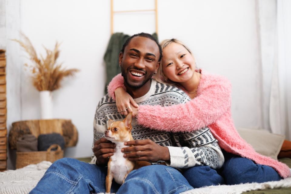 Smiling young diverse couple looking at camera and hugging near pet sitting on their bed