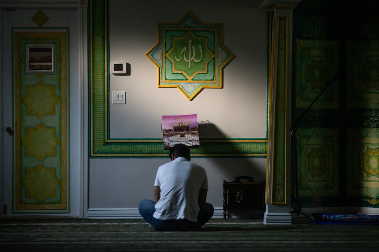 A man prays in a dimly lit mosque as a beam of sunlight casts along a wall.