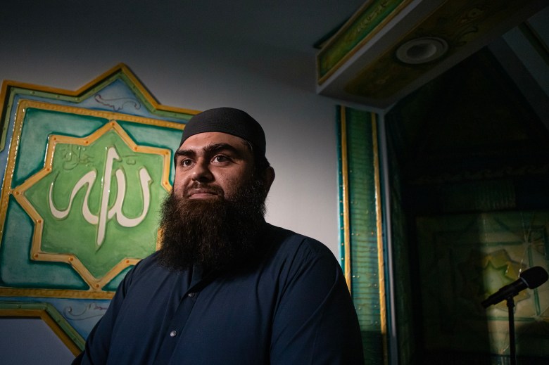 Imam Qarim Abdullah stands near a sign with Arabic lettering inside a mosque, as a beam of sunlight is cast on the wall.