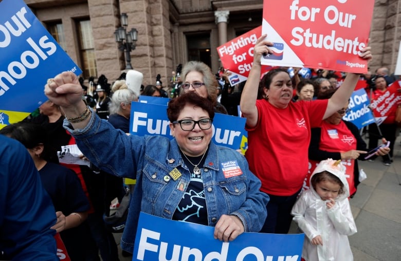 A woman with short hair, glasses and a denim jacket stands amid a group of protesters, pumping one fist in the air and holding a sign with her other hand that reads 