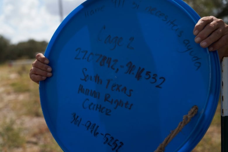 A pair of hands holds up a big blue garbage bin lid with the name and phone number for the South Texas Human Rights Center scrawled on it in black marker.