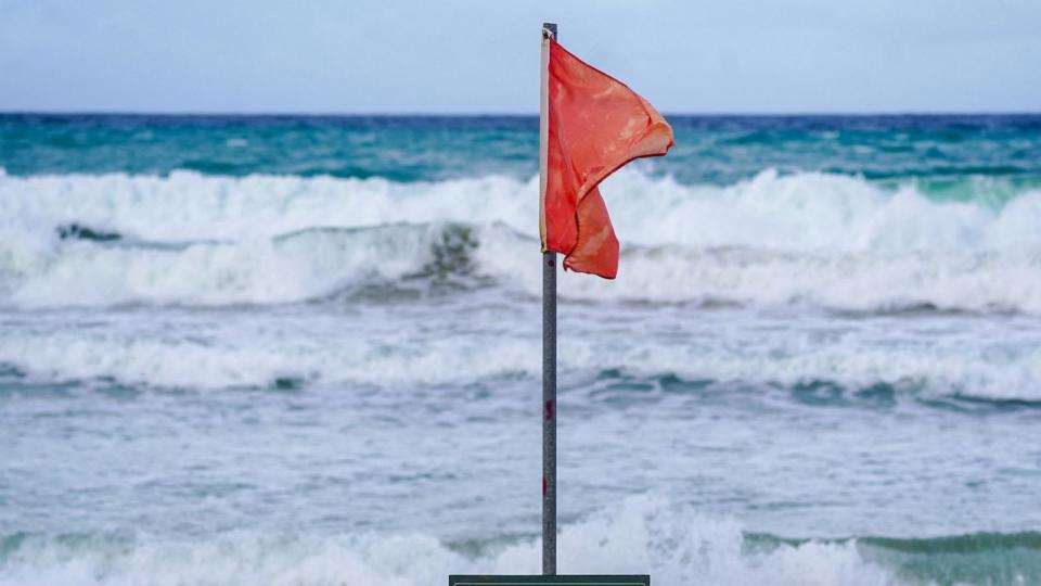 PHOTO: A warning sign reading 'Danger, No Swimming' under a red flag, indicating unsafe swimming conditions, are seen on the beach as waves crash onto the shore in Luquillo, Puerto Rico, on August 13, 2024, as Tropical Storm Ernesto approaches.  (Jaydee Lee Serrano/AFP via Getty Images)