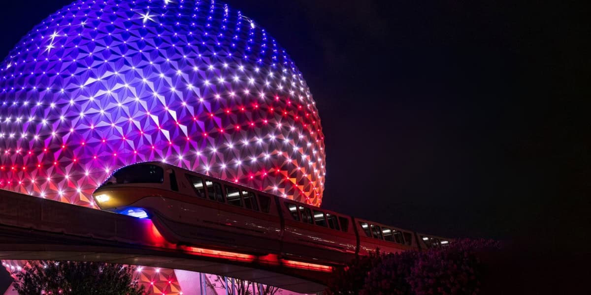 A monorail passes in front of a large, spherical structure lit up with a gradient of red, white, and blue lights at night, creating a futuristic atmosphere. The structure's geometric pattern is highlighted by the lighting. Trees are visible below the platform.