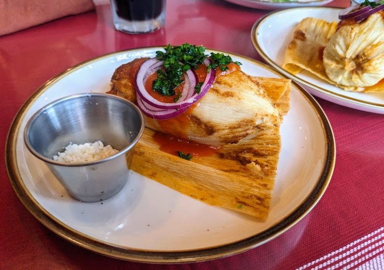 A plate of tamale garnished with red onion and cilantro, served with a side of white cheese in a small metal cup on a red tablecloth.