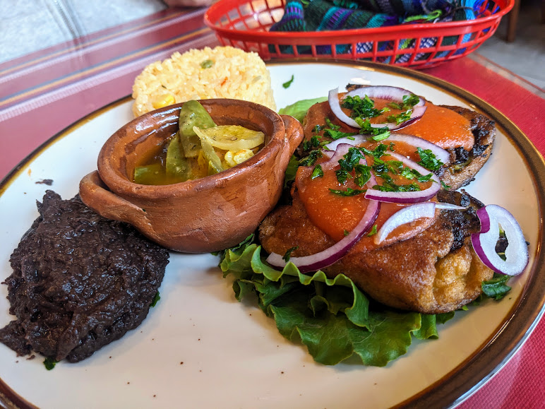 A plate of Mexican food featuring two stuffed peppers topped with sauce and onions, accompanied by black beans, yellow rice, and a small dish of cooked vegetables.