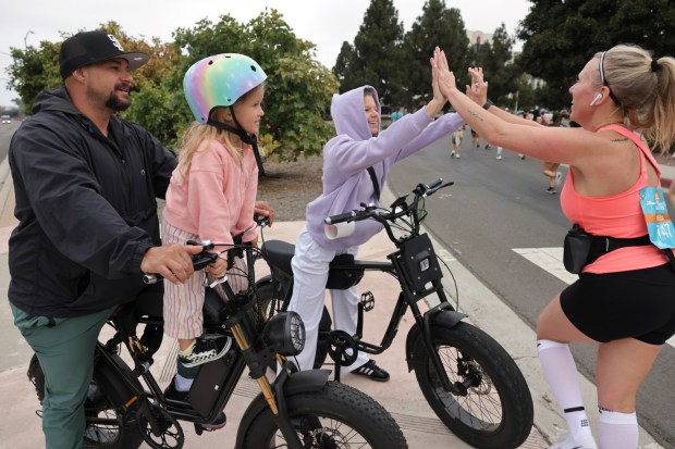 John Hodge and daughter Tatum, 5, watch as his wife Vineca Hodge high fives daughter Marlee, 13, who are following on e-bikes, as she and thousands of people participating in the half marathon run on Harbor Island during the 47th annual America's Finest City Half Marathon and 5K in San Diego on Sunday, Aug. 18, 2024. Over 1,300 people participated in the 5K and more than 4,300 in the half marathon. (Hayne Palmour IV / For The San Diego Union-Tribune)