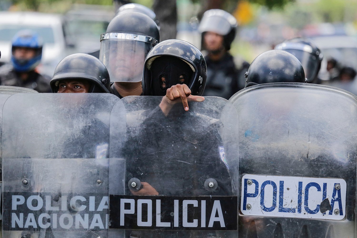 The helmet-wearing heads of a riot police force are visible above their scuffed riot shields. At the center, one of the officers wearing a mask points over his shield at the photographer.