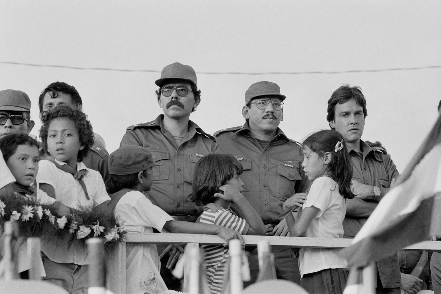 In this black-and-white photo from 1982, Daniel Ortega and his brother Humberto Ortega are seen in military uniform and glasses as they watch a military parade. Other FSLN leaders stand around them, as do a crowd of children.