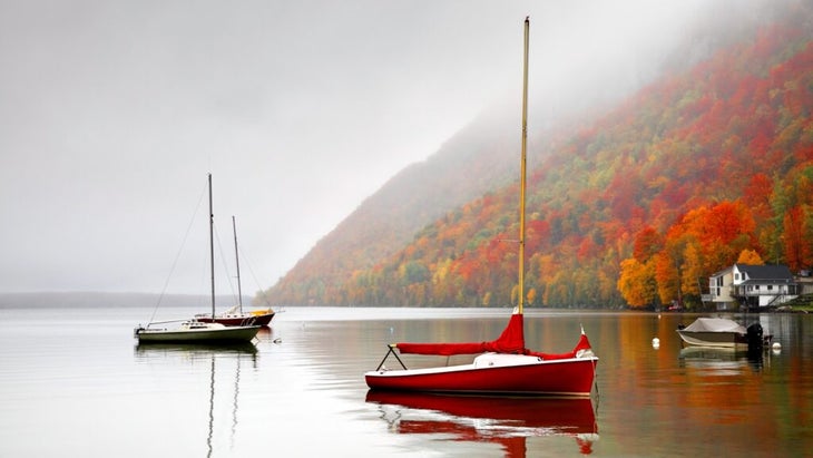 Boats are moored on Lake Willoughby, Vermont. It's a foggy day and the steep hillsides are covered in trees at the peak of fall foliage.