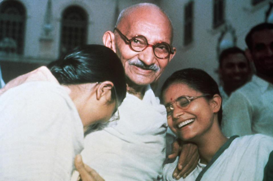 Mahatma Gandhi is smiling and embracing two women who are laughing and wearing traditional attire