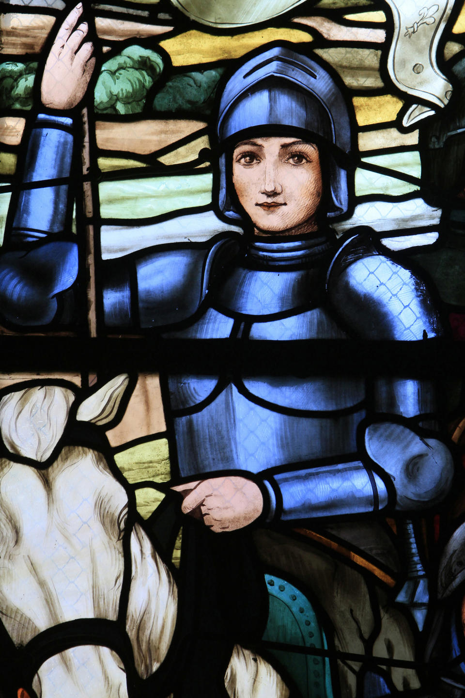 Stained glass artwork depicting Joan of Arc in armor, holding a sword and riding a horse. Medieval landscape background