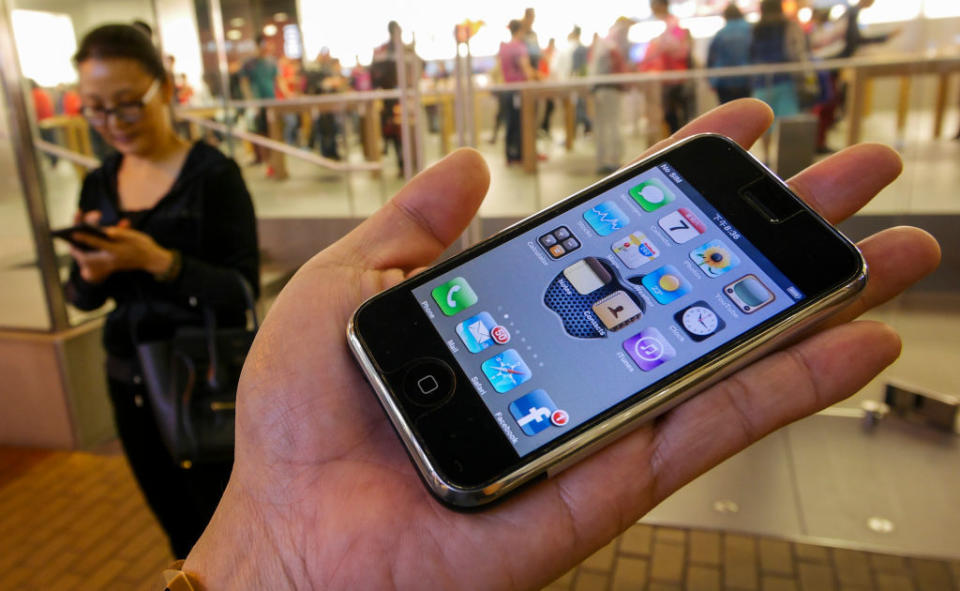 A person holds an early model iPhone displaying apps on the screen. In the background, another person stands using a phone