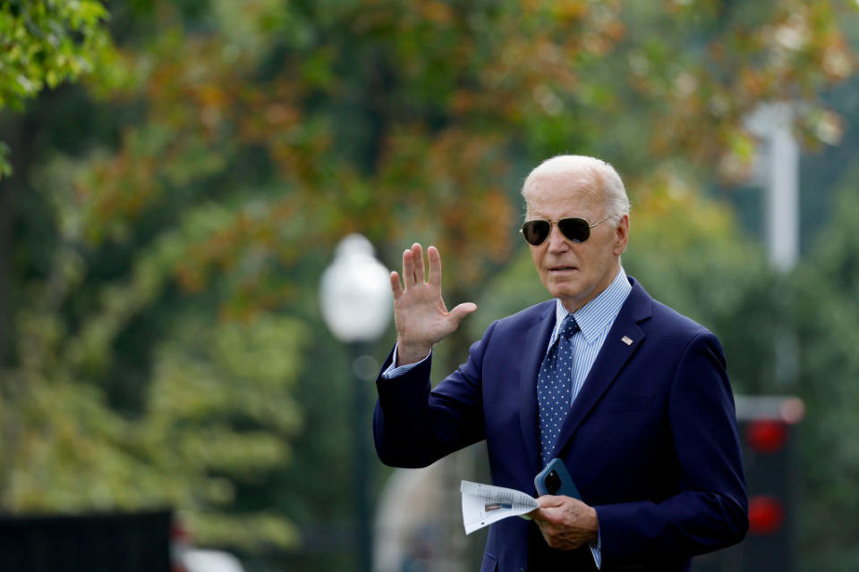 Joe Biden, wearing a suit and sunglasses, waves while holding a piece of paper and a phone. Trees and blurred background visible