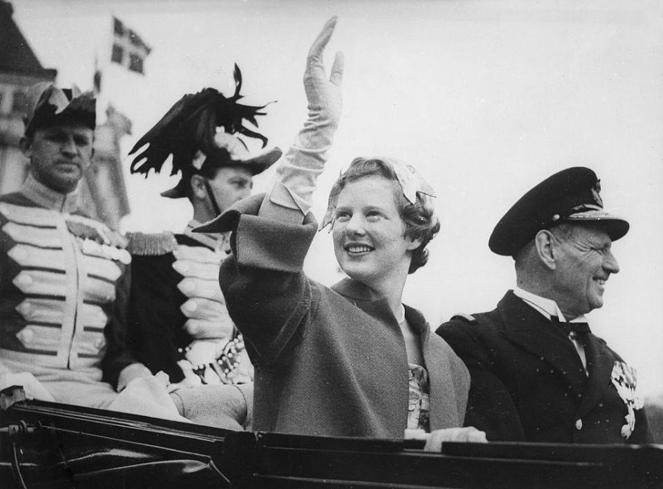 Margaret Thatcher waves to a crowd while aboard a vehicle. She is dressed in formal attire, wearing gloves. Accompanying her are military personnel