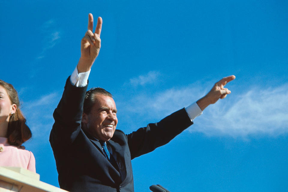 Richard Nixon smiling, raising both arms and making V-signs with his fingers. An unidentified person is partially visible on the left side