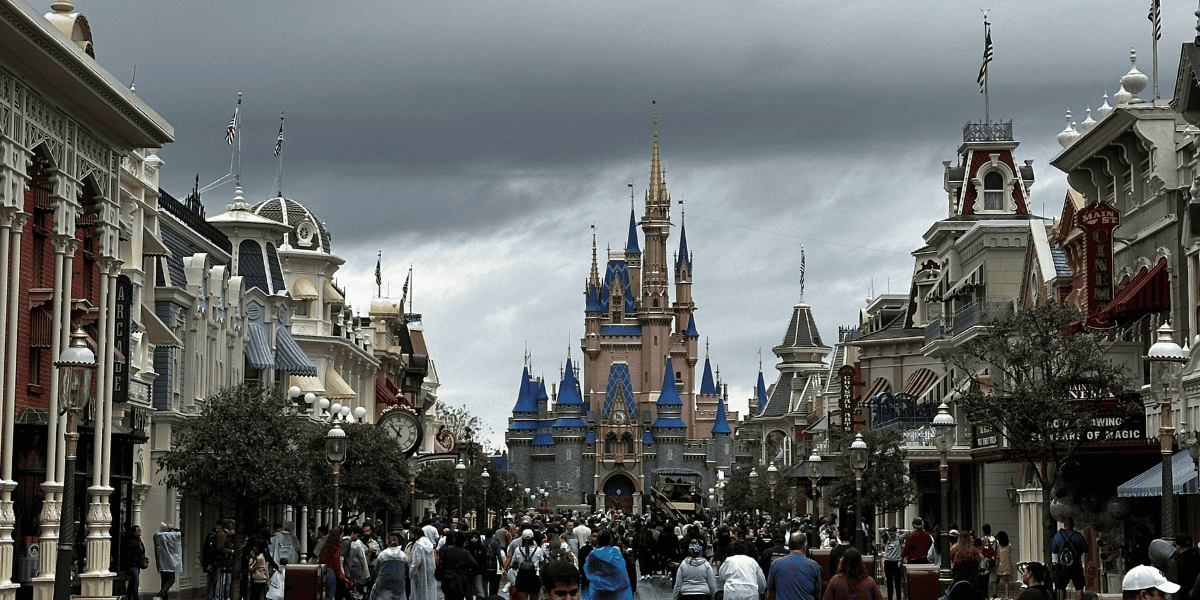 A bustling main street lined with picturesque buildings under a moody sky, leading to a Disney castle in the distance, filled with visitors exploring the enchanting surroundings under watchful security.