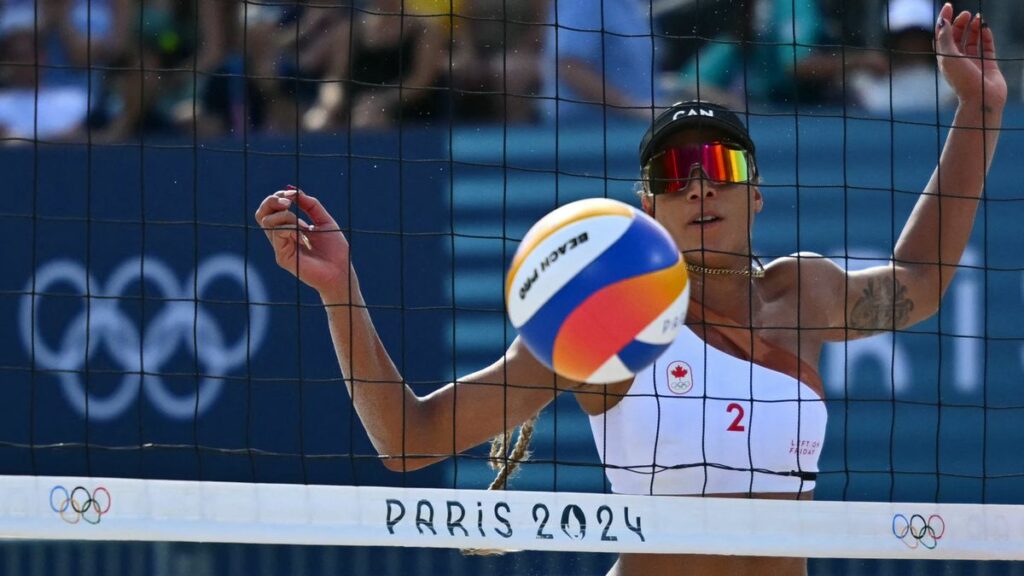 Brandie Wilkerson of Canada, with a white bikini top, black visor and mirrored sunglasses, eyes the ball through the net at the 2024 Olympic Beach Volleyball.