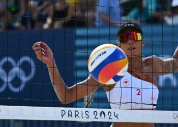 Brandie Wilkerson of Canada, with a white bikini top, black visor and mirrored sunglasses, eyes the ball through the net at the 2024 Olympic Beach Volleyball.