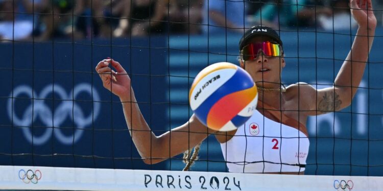 Brandie Wilkerson of Canada, with a white bikini top, black visor and mirrored sunglasses, eyes the ball through the net at the 2024 Olympic Beach Volleyball.