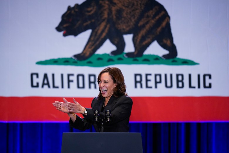 Vice President Kamala Harris stands while clapping behind a podium and in front of a large California state flag.