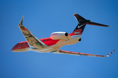 A small jet in flight with a red and white body, a black tail with a white Nike Swoosh logo and colorful mural-like decoration under the wings