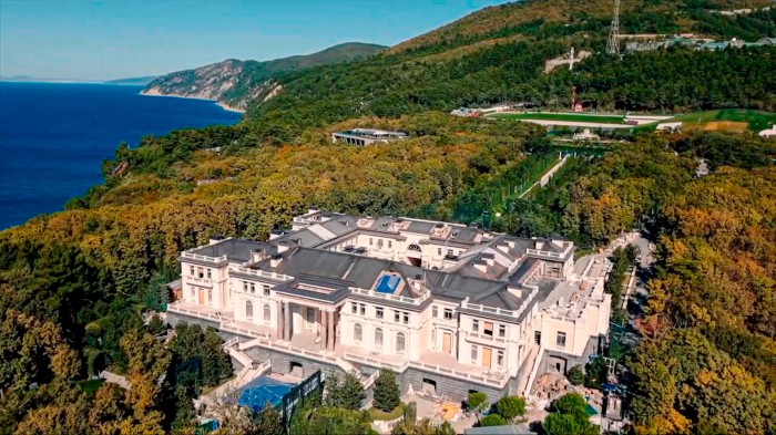 An aerial view of a very large white building surrounded by trees, with the sea in the distance