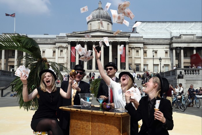 Five smiling people in bowler hats sit or stand at a table where there is a cocktail glass. All are holding notes, one person is throwing them up in the air 