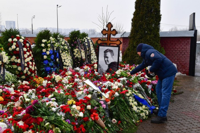 A figure in a blue puffer jacket and woolly hat leans down to add a bunch of flowers to the many floral tributes surrounding a large photograph of Alexei Navalny