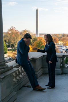 A dark-haired man laughs into his hands as a dark-haired woman smiles in front of him. The needle-like Washington Monument is in the background.