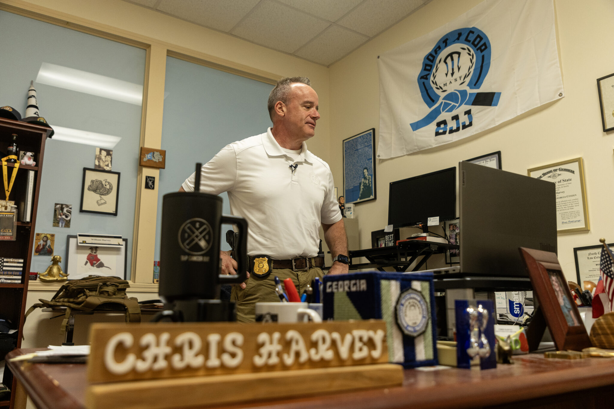 A white man with gray hair in a white polo shirt with a sheriff's badge stands in his office.