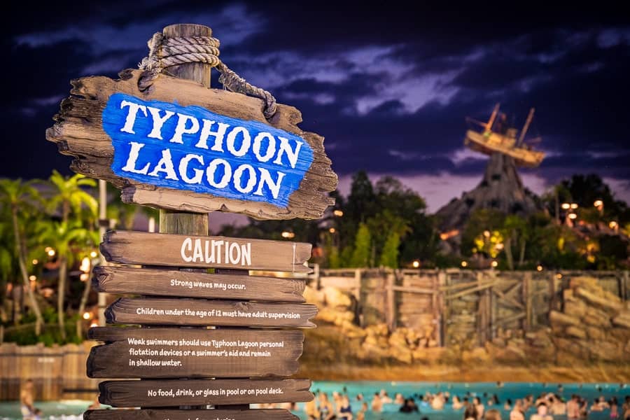 A sign at Typhoon Lagoon warns of strong waves and safety precautions, while guests swim in the pool with a shipwrecked boat in the background at dusk.