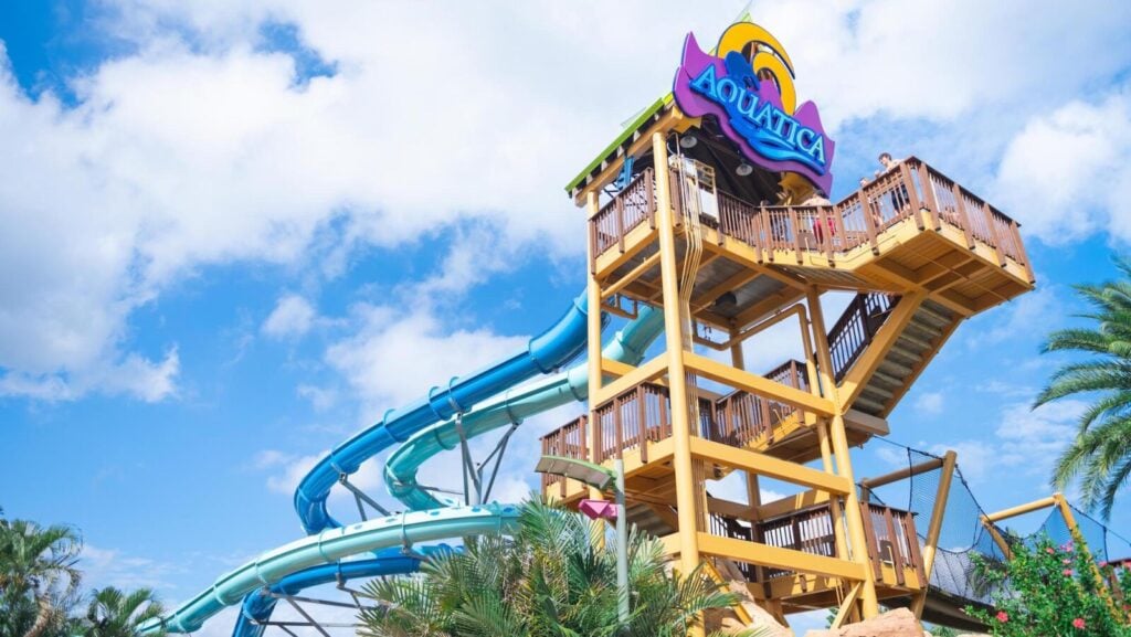 A tall waterslide structure at Aquatica theme park against a blue sky with clouds and tropical plants in the foreground.