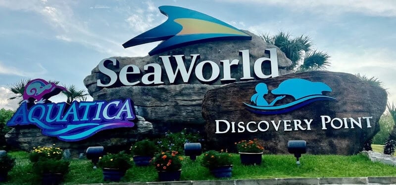 Entrance sign for SeaWorld, Aquatica, and Discovery Point featuring logos and surrounded by potted plants and greenery.