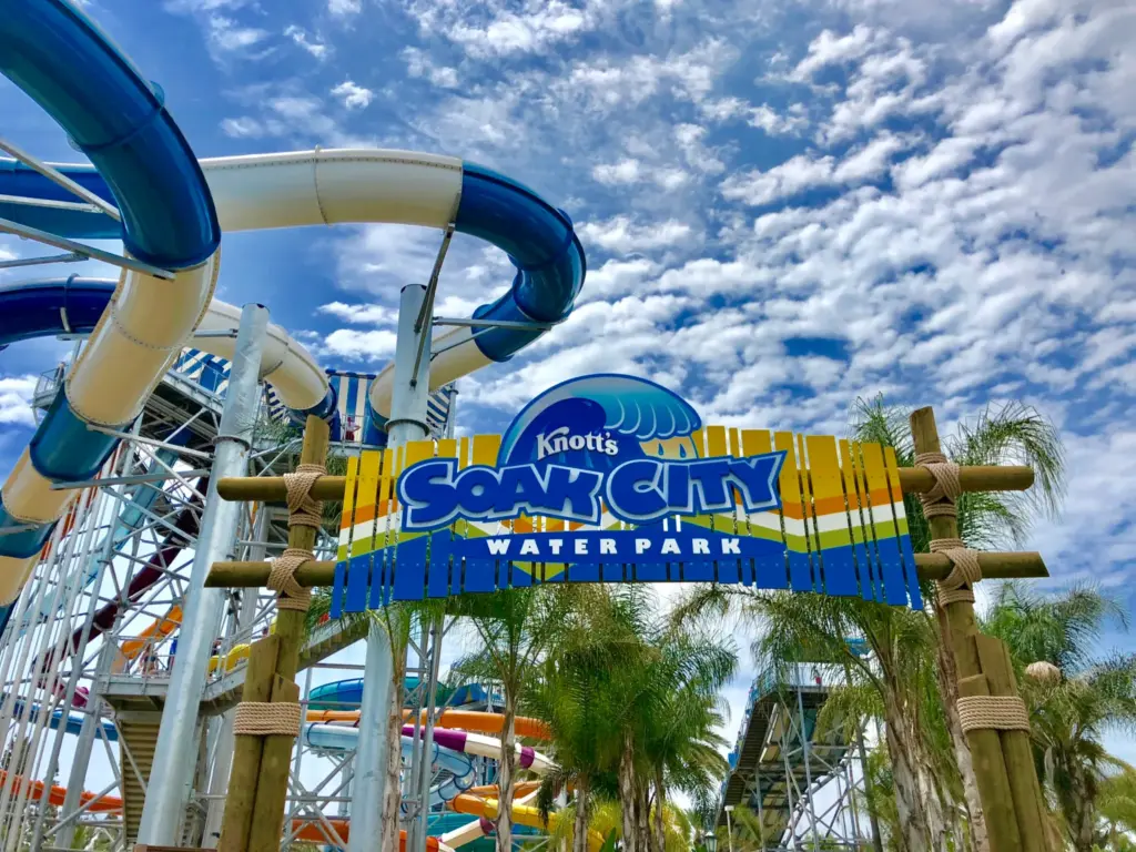 Entrance sign for Knott's Soak City Water Park, with water slides and palm trees in the background under a partly cloudy sky.