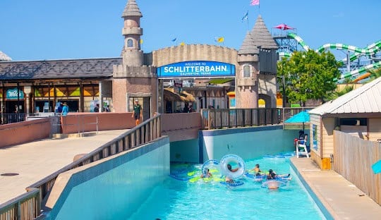 People float on inner tubes in a lazy river at Schlitterbahn Waterpark in Galveston, Texas, with a castle-themed entrance in the background.