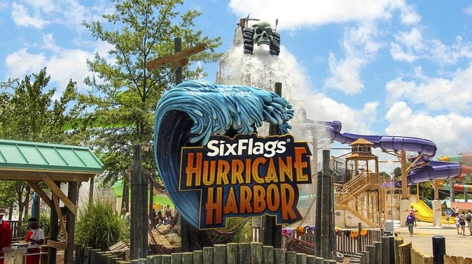 Entrance sign for Six Flags Hurricane Harbor with a water ride and colorful slides visible in the background under a partly cloudy sky.