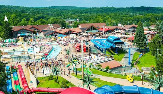 Aerial view of a busy water park with various slides and attractions, surrounded by greenery and buildings. People are enjoying different activities, and the weather appears to be sunny and clear.